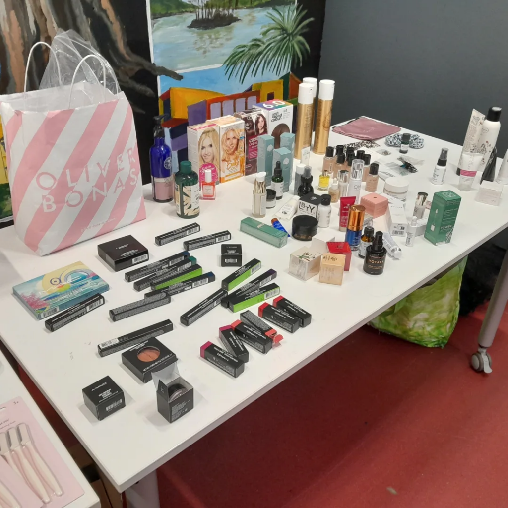 Assorted makeup, skincare and hair products laid out on a white table with bright coloured artwork behind.