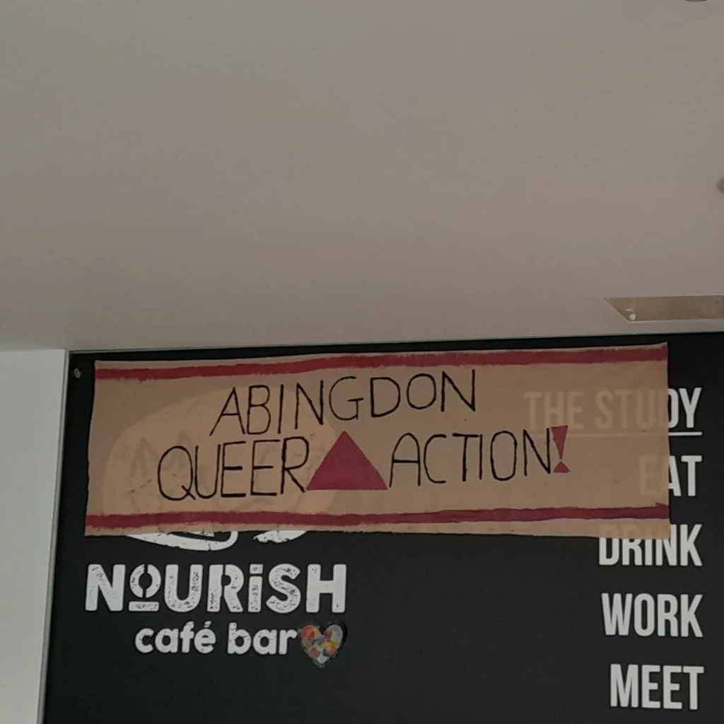 Pink and black Abindon Queer Action banner with a pink triangle in the middle, hung over a sign for the "Nourish Cafe Bar".