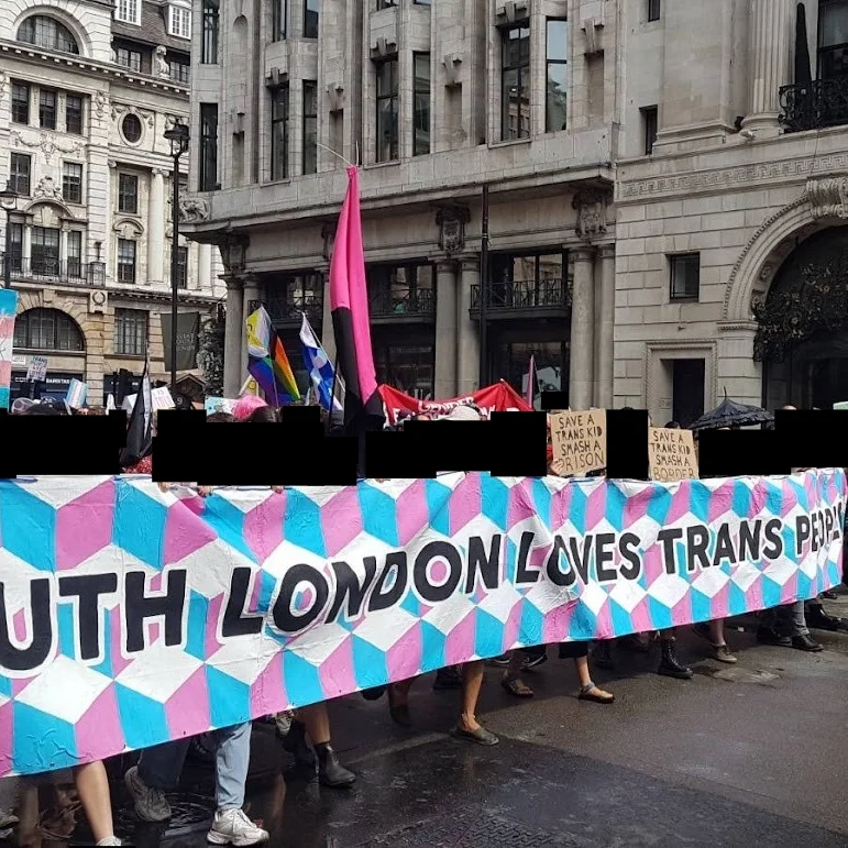 Image of a crowd of people marching down a road. People in the photo are holding a pink, white and blue banner which says "South London loves trans people". There are other signs in the photo which say: "Let trans people bloom", "Save a trans kid smash a border", "Save a trans kid smash a prison". There are also flags in the photo. The faces and identifying features of the crowd have been edited out with black squares.
