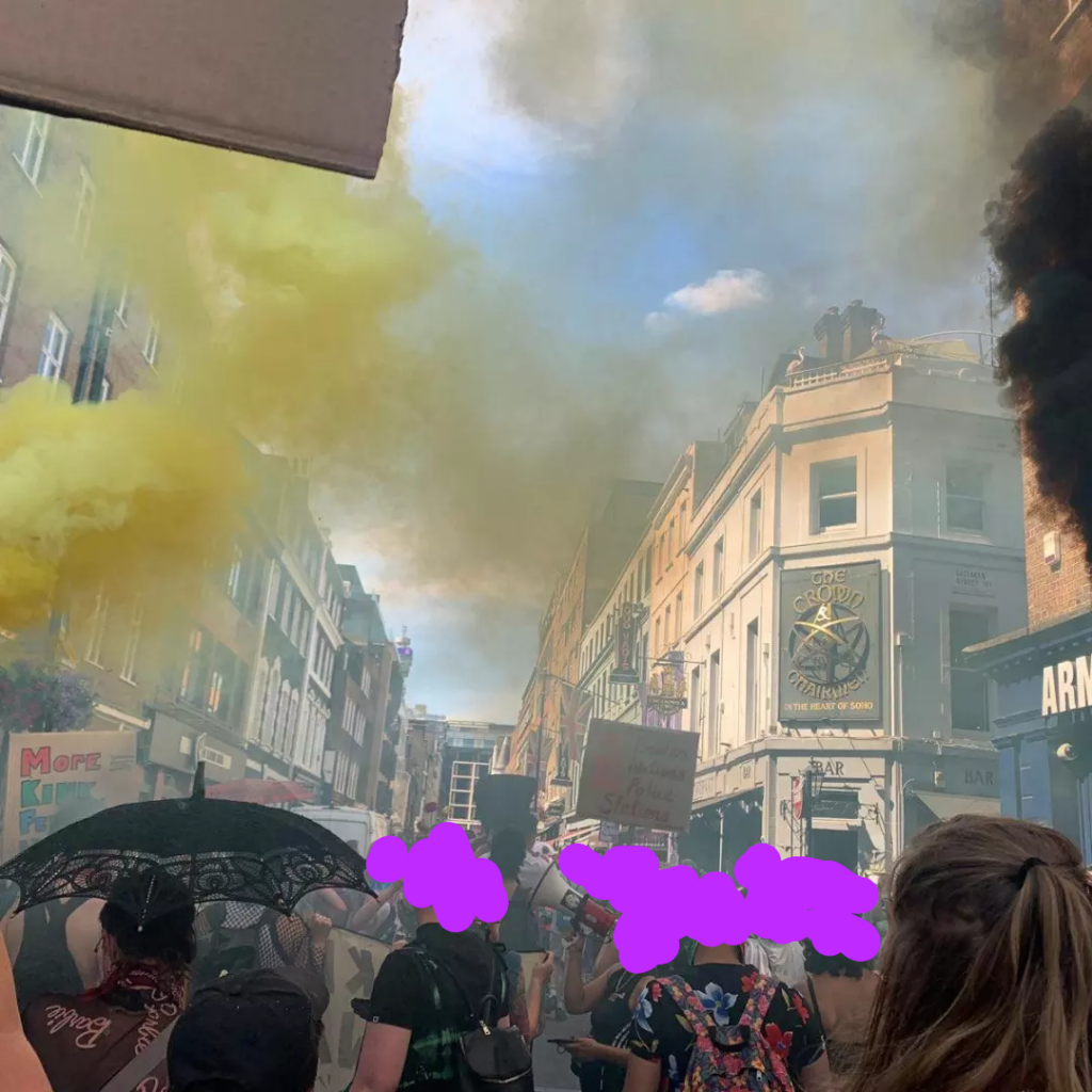 A London street filled with people featuring yellow and black smoke from flares across the photo. There is also a sign in the photo that says “No borders, No nations, No police stations.”