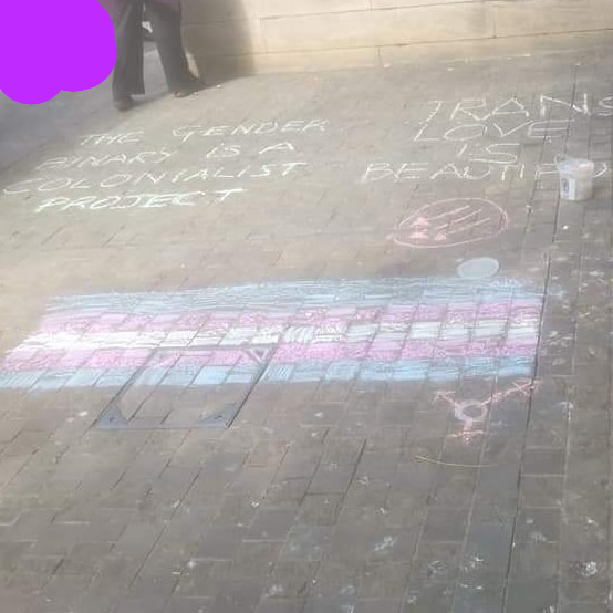 Coloured chalk on the ground including a trans flag and "trans love is beautiful" and "the gender binary is a colonialist project"