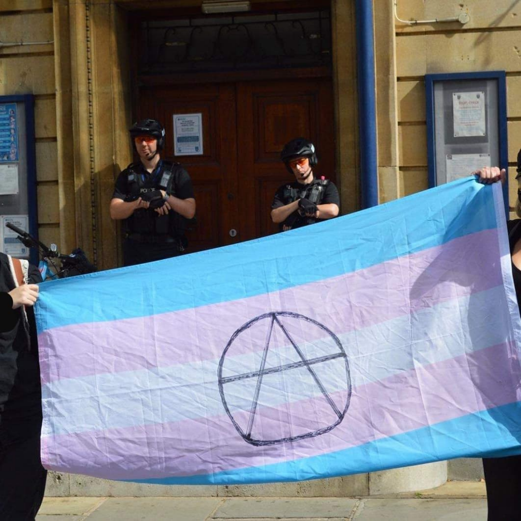 a trans anarchist flag being held in front of 2 police officers standing in front of the doors to a police station