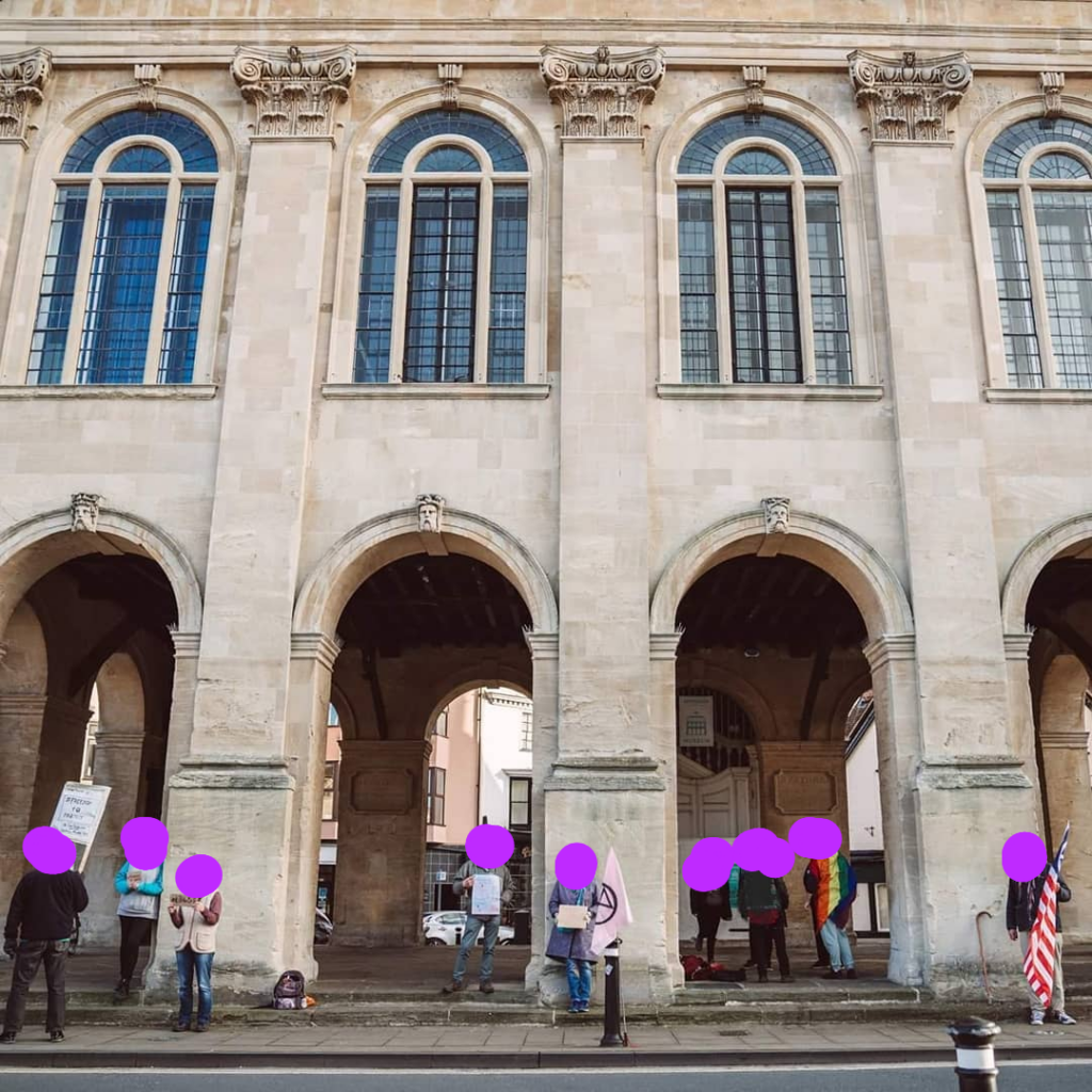 A group of people spread out standing in front of an old stone building with signs and flags