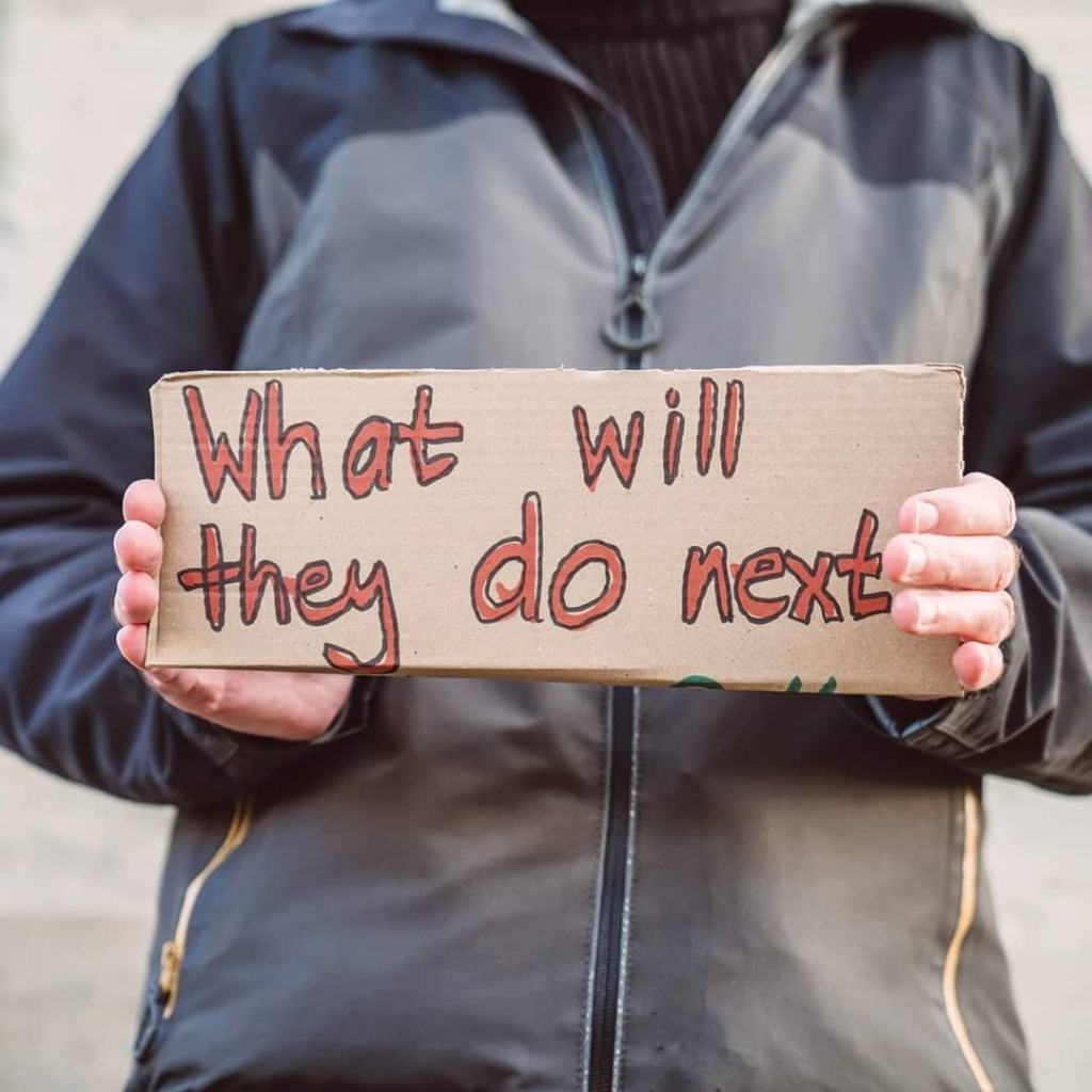 Person in a grey coat holding a cardboard handwritten sign that says "What will they do next" in red