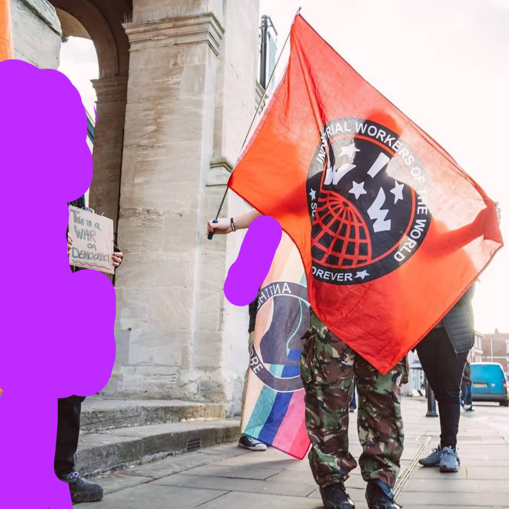 From left to right, a person holding a cardboard sign that says "this is a war on democracy", a person holding a red IWW flag and another person holding a rainbow anti homophobe action flag. In front of a stone pillar on the street