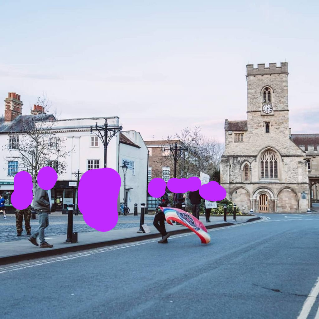 Various people spread out around a market square with one person in the middle holding a rainbow flag and others holding signs