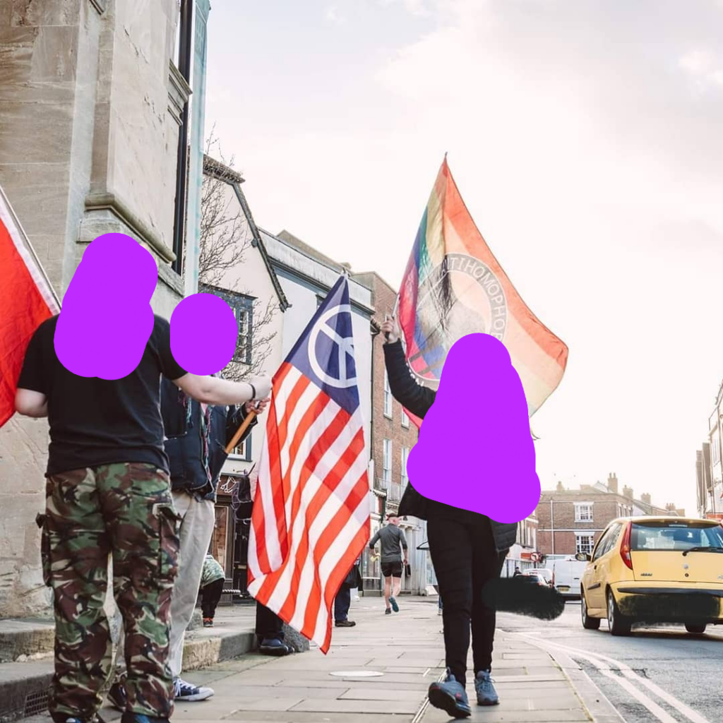 People holding different flags on the street including a american peace flag and an anti homophobe action rainbow flag