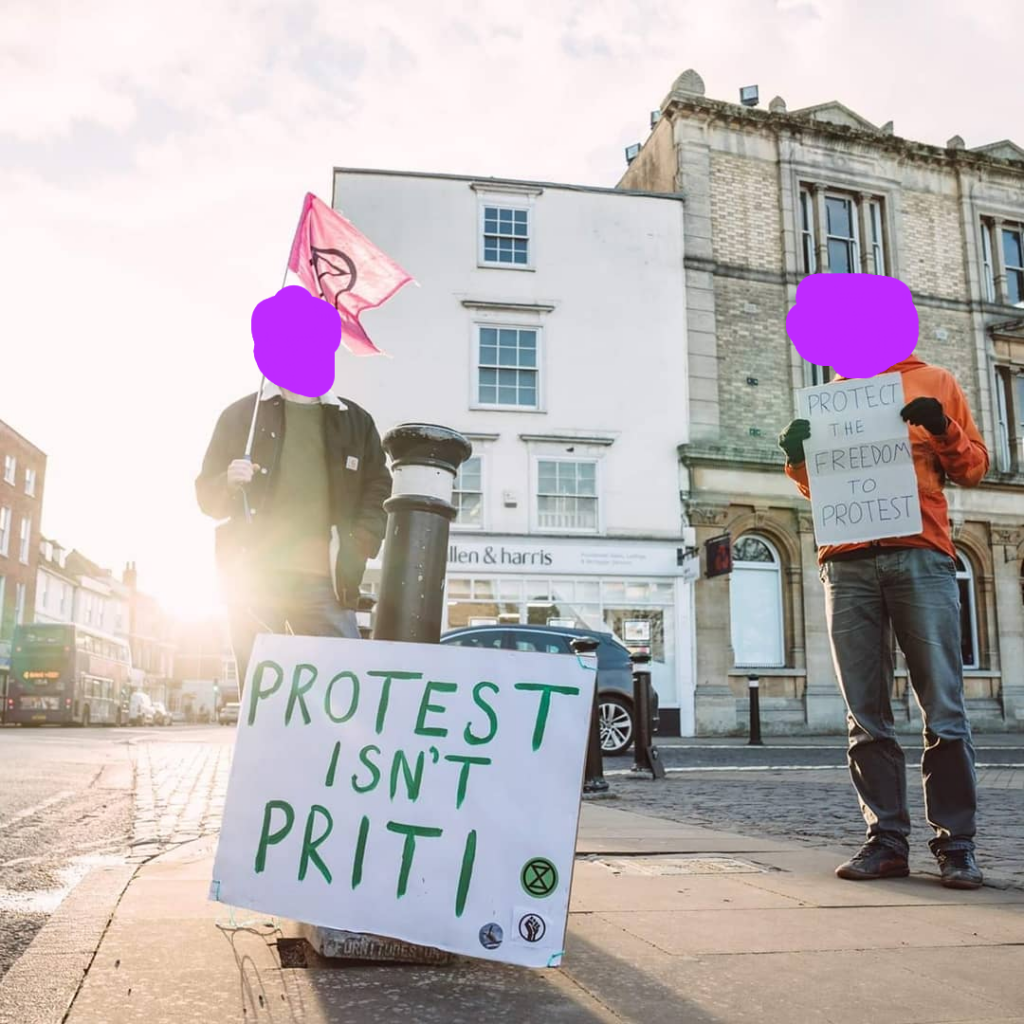 a person holding an extinction rebellion flag with a sign that says "Protest isn't Priti" next to a person holding a sign that says "Protect the freedom to protest"