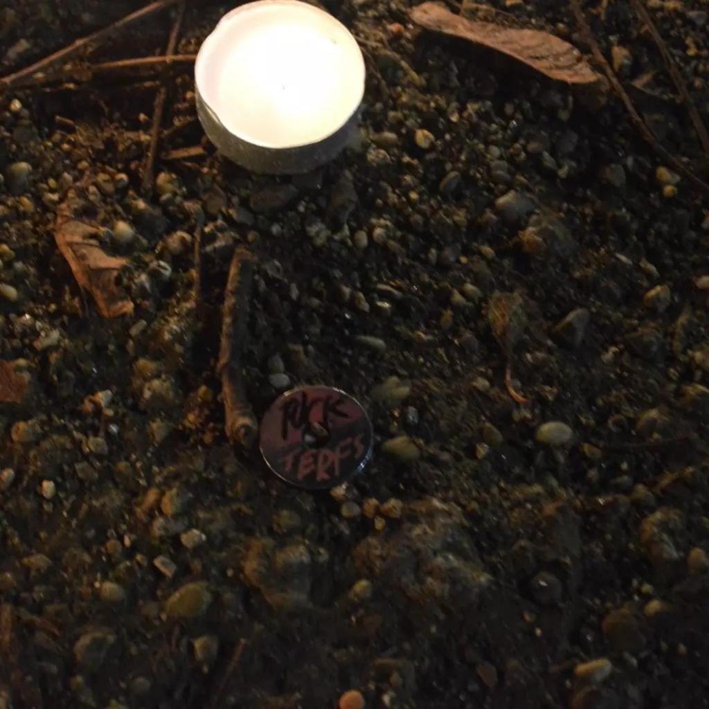 A close up of a tealight and a pin that says “fuck terfs”.