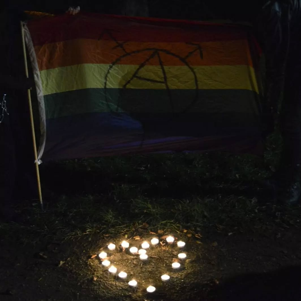 Two people holding gay pride flag (with the tranarchy symbol on it) and a tealight heart shape on the ground.
