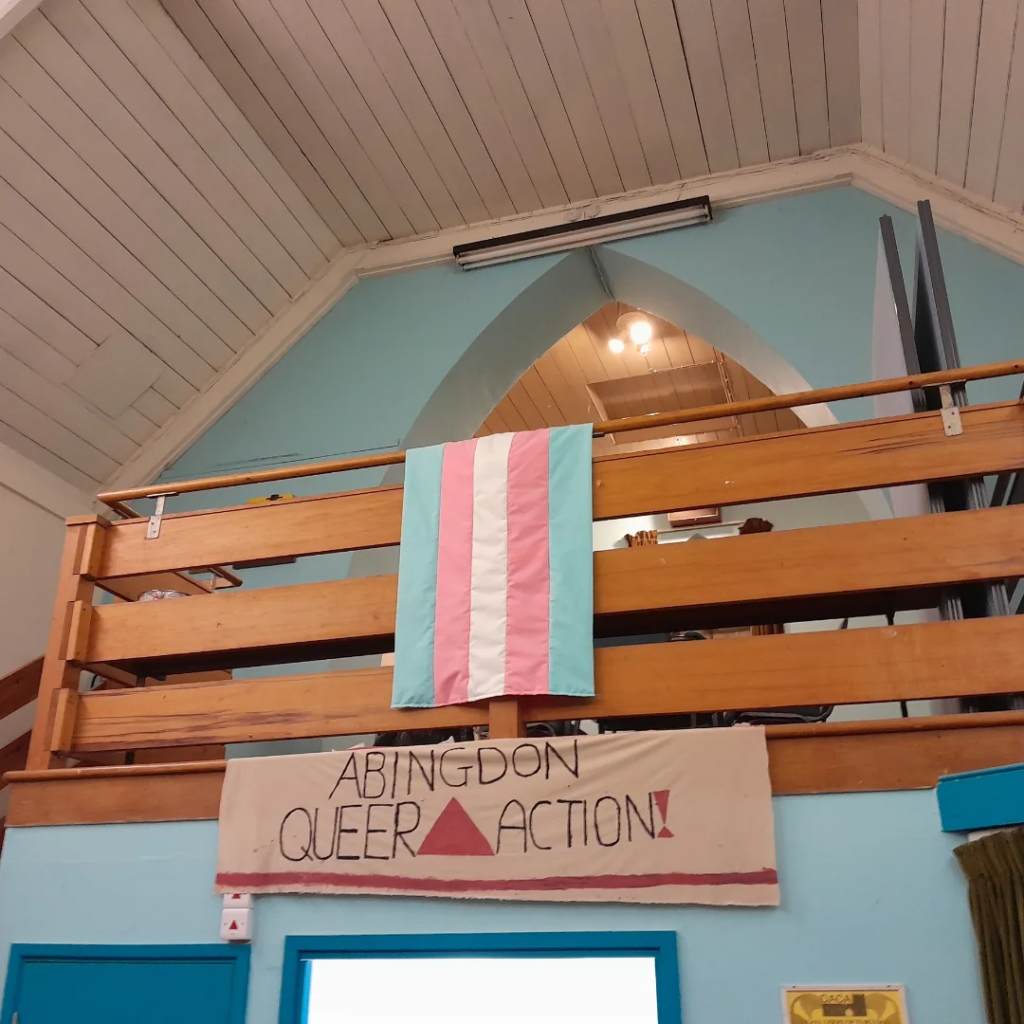 Trans flag hanging vertically from a wooden railing above a beige and pink Abingdon Queer Action banner.