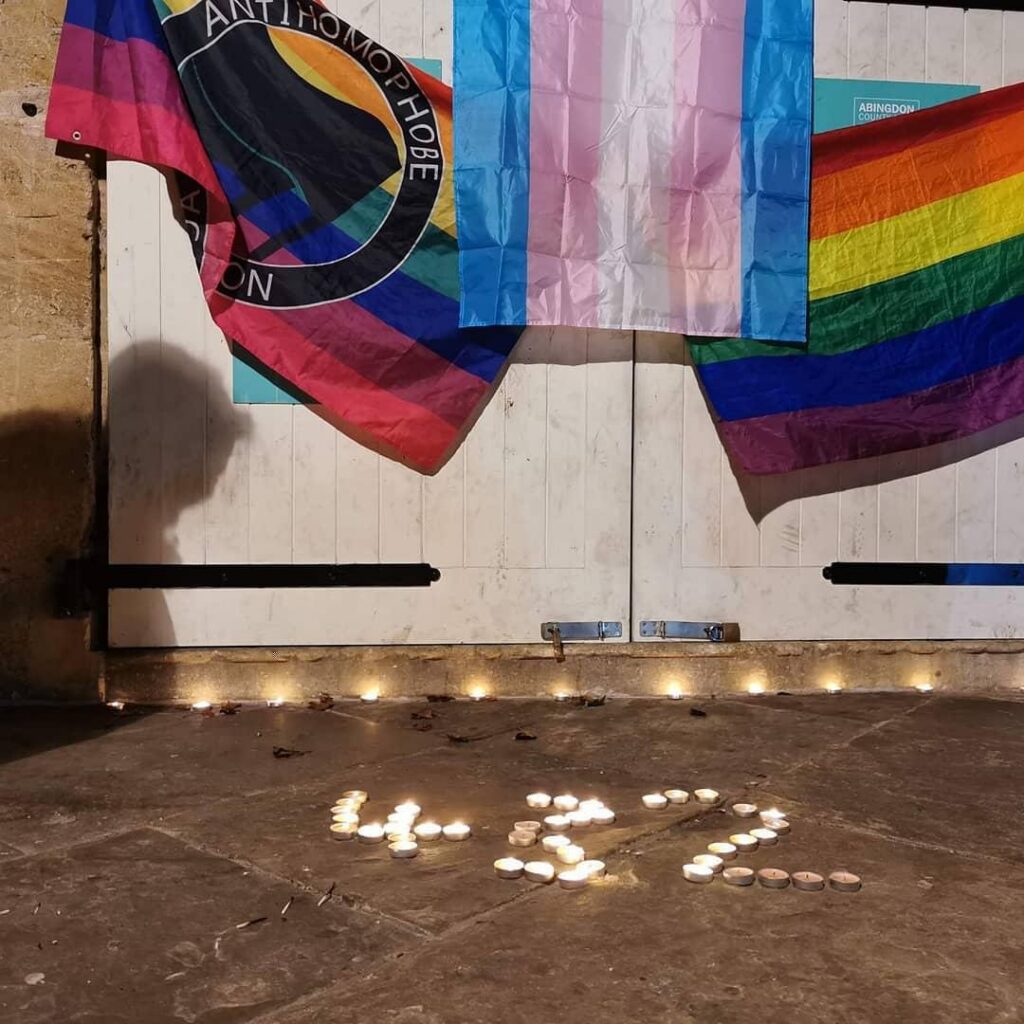 an anti homophobe action flag, a transgender flag and a rainbow flag hanging on white doors with the numbers 432 made using candles on the floor below
