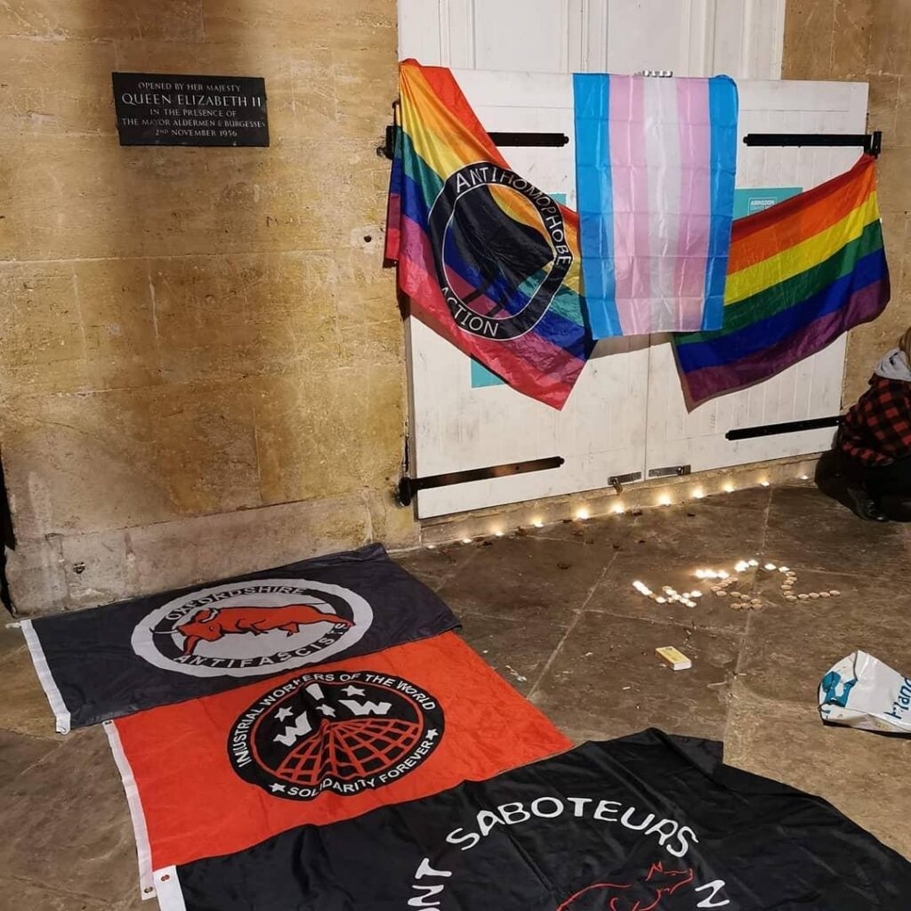 an anti homophobe action flag, a transgender flag and a rainbow flag hanging on white doors with the numbers 432 made on the floor in front and an oxfordshire antifascists flag, an IWW flag and a hunt saboteurs flag on the floor next to the candles
