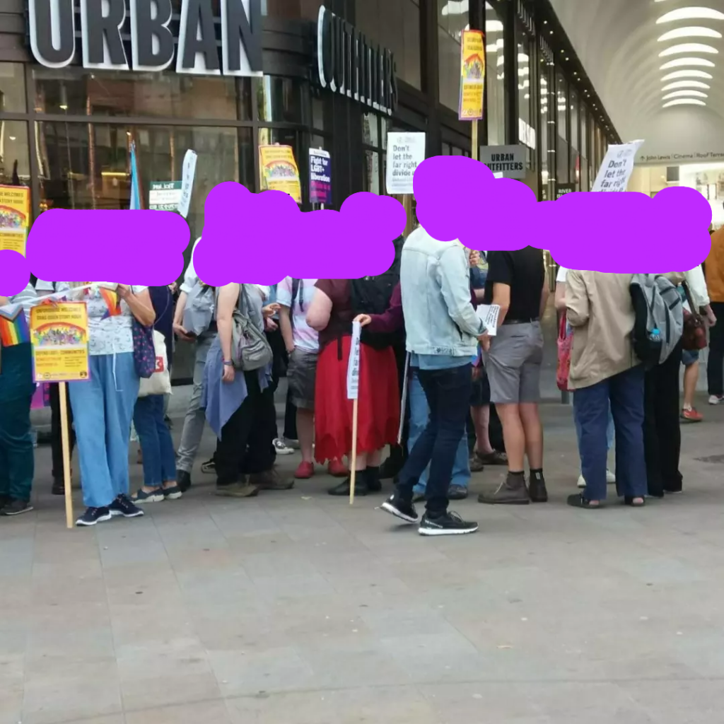 Group of people standing in front of a shopping centre entrance with pro queer signs and flags