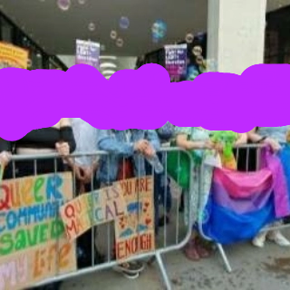 A group of people standing behind crowd barriers with signs including "Queer community saved my life" and "Queer is magical" and "You are enough" also queer flags