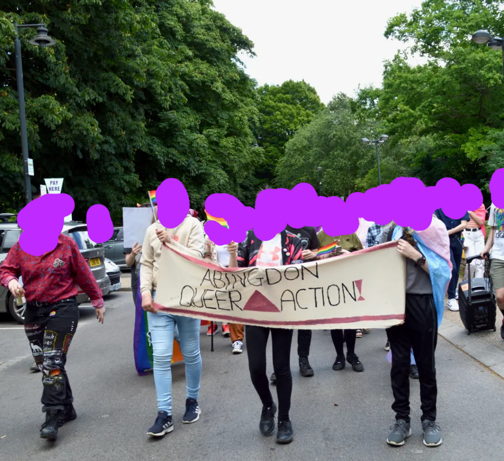 The front of the pride parade with people holding a abingdon queer action banner and queer flags walking down the street