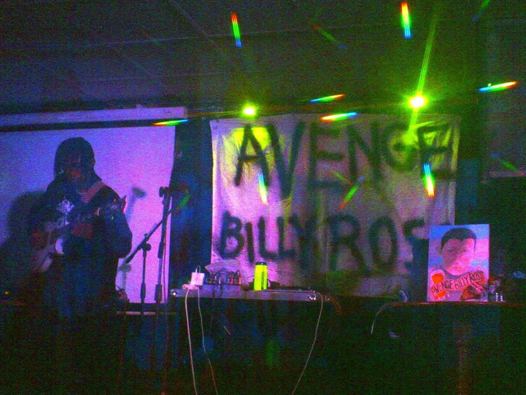 Person on a stage playing a guitar with yellow lights behind them around a banner saying "Avenge Billy Ross" - to the right is a painting of billy on a trans flag background.