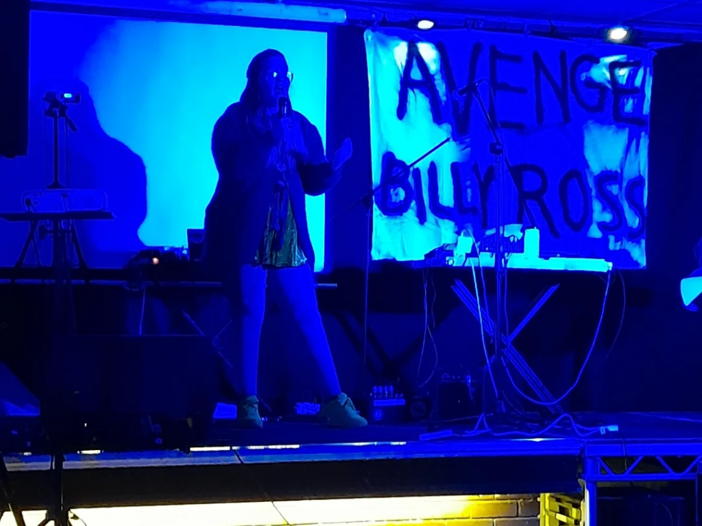 Person on a stage singing with a microphone lit by blue lights with a banner saying "Avenge Billy Ross" behind them.