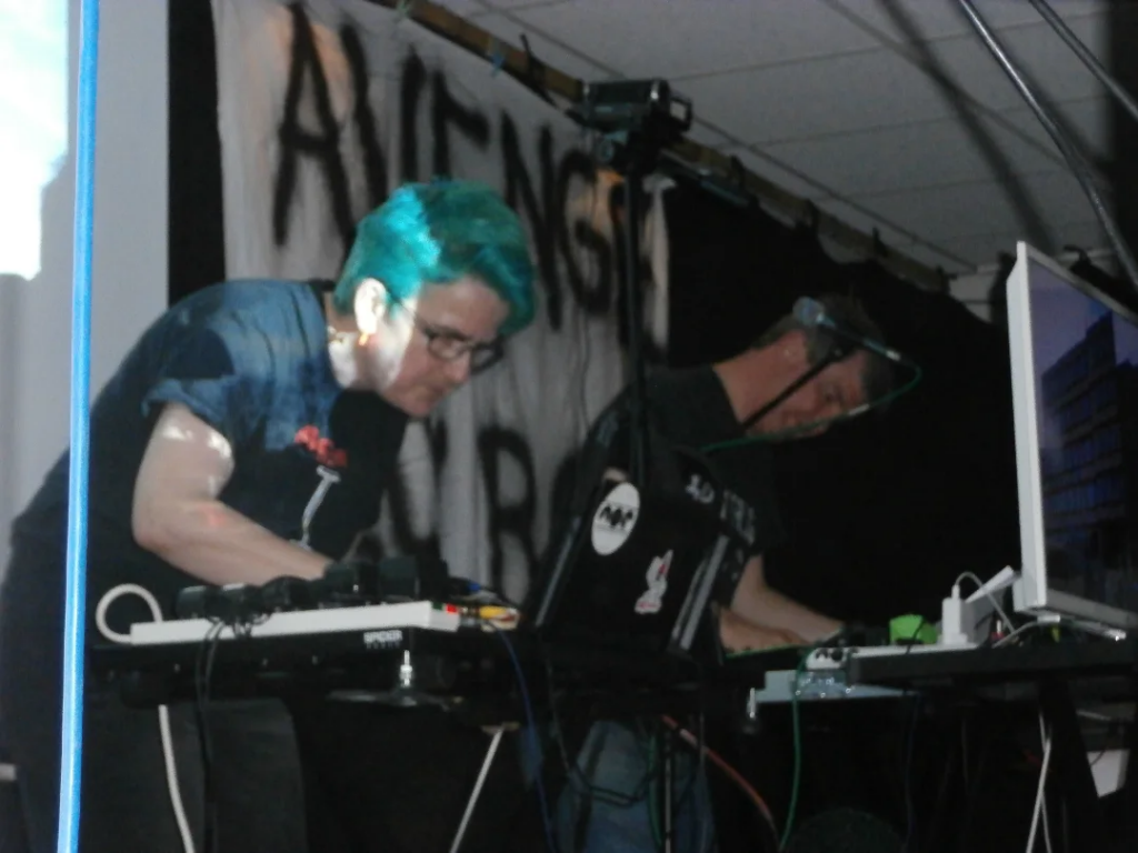 Two people leaning over keyboards on a stage with monitor in front of them.
