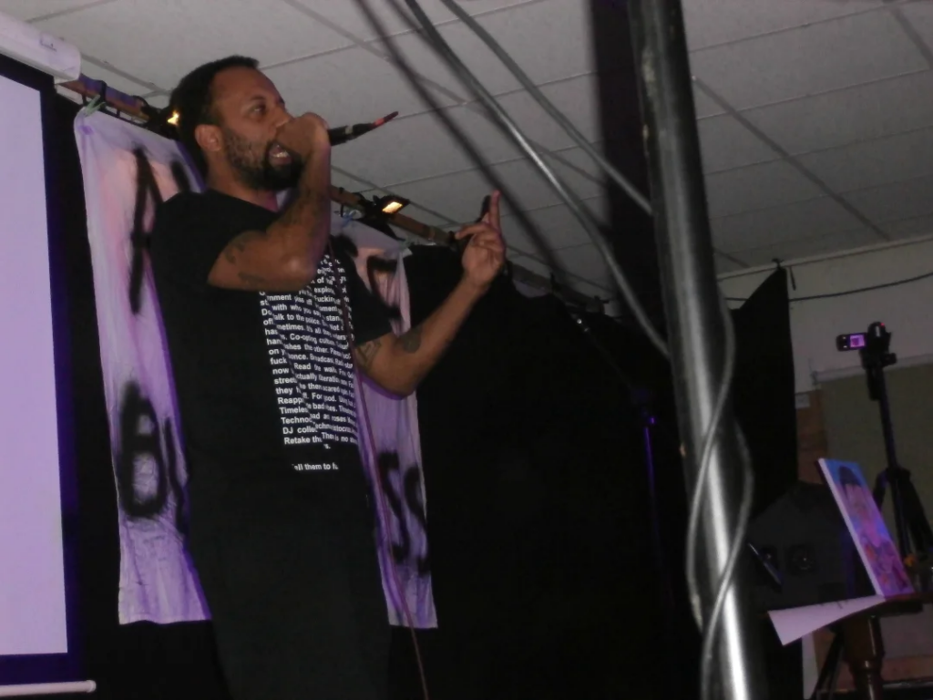 Person on a stage gesturing and holding a microphone.