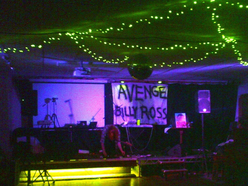 Person crouched over on a stage lit by purple lighting, with yellow fairy lights suspended from the ceiling.