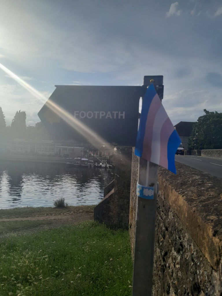 Small trans flag attached to a sign which says "Footpath". The river and road is visible behind the sign and there is a ray of sunlight in the forefront of the image.